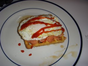 Waffle layered with maple syrup, smoked salmon, a sunnyside up egg, ketchup, and sriracha